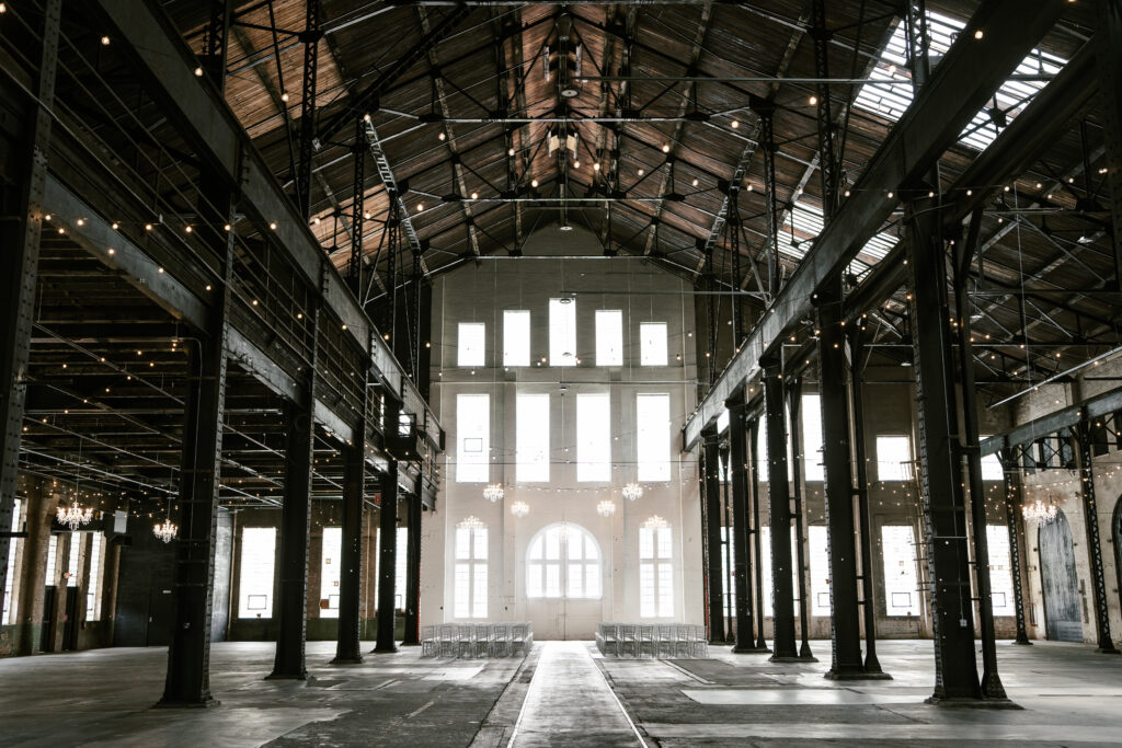 Picture of venue with high ceilings, black beans, and a white wall. Has alot of windows and string lights to light up the space